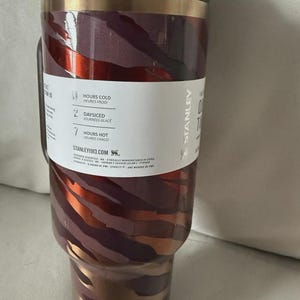 May include: A Stanley insulated tumbler with a handle. The tumbler features a maroon, copper, and gold zebra stripe design. The lid and handle are dark brown. The tumbler has a white label with text indicating it keeps drinks cold for hours.