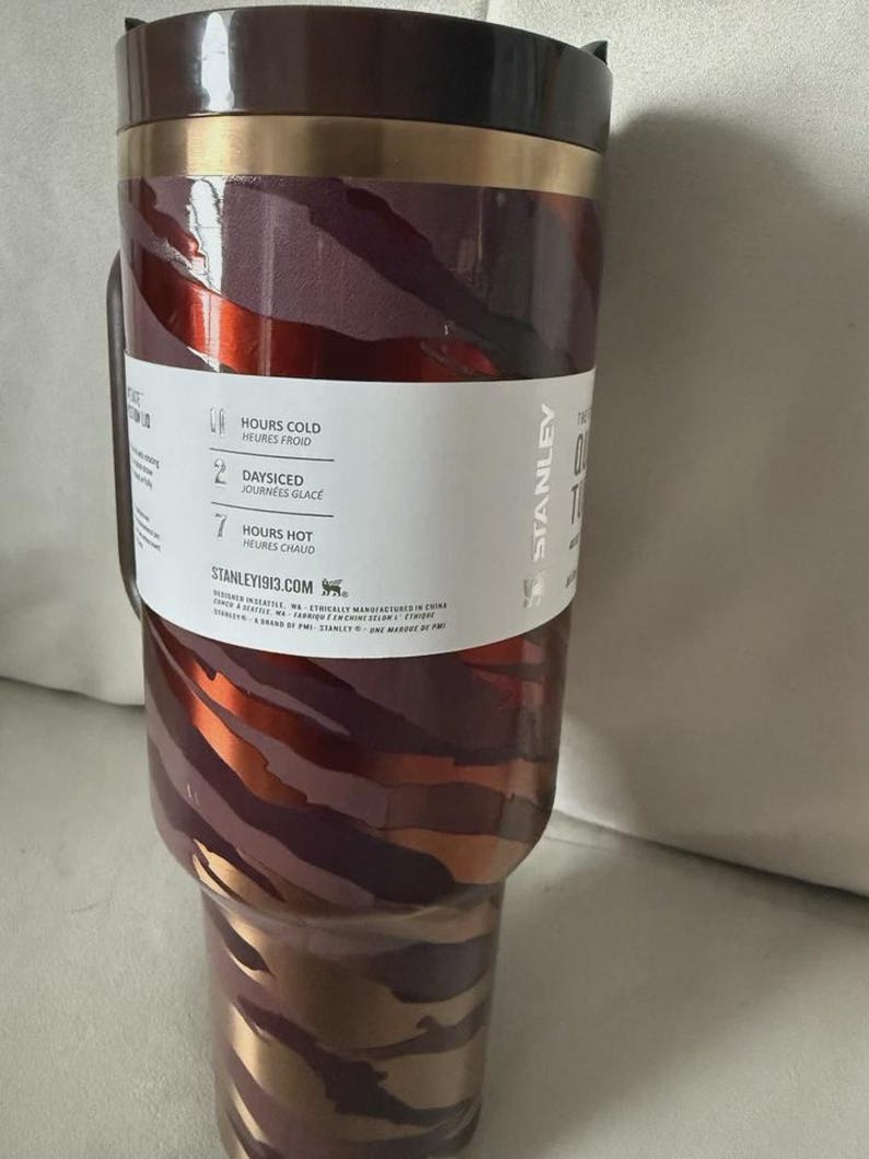 May include: A Stanley insulated tumbler with a handle. The tumbler features a maroon, copper, and gold zebra stripe design. The lid and handle are dark brown. The tumbler has a white label with text indicating it keeps drinks cold for hours.