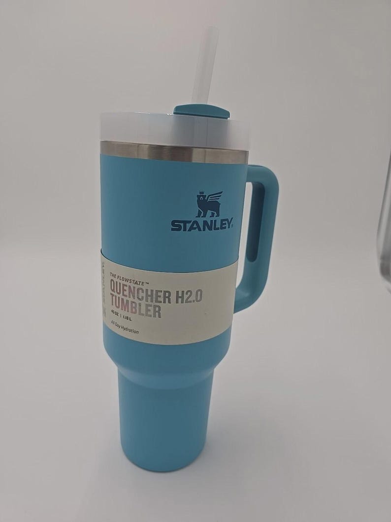 May include: A light blue Stanley Quencher H2.0 tumbler with a handle and straw. The tumbler has a white band with the text "QUENCHER H2.0 TUMBLER" and the Stanley logo. It holds 1.18L and is designed for all-day hydration.