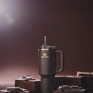 Peut inclure: Un mug de voyage Stanley noir avec une poign&eacute;e et une paille. Le mug a un bord dor&eacute; et le logo Stanley en or. Le mug est pos&eacute; sur une surface sombre avec des morceaux de chocolat.