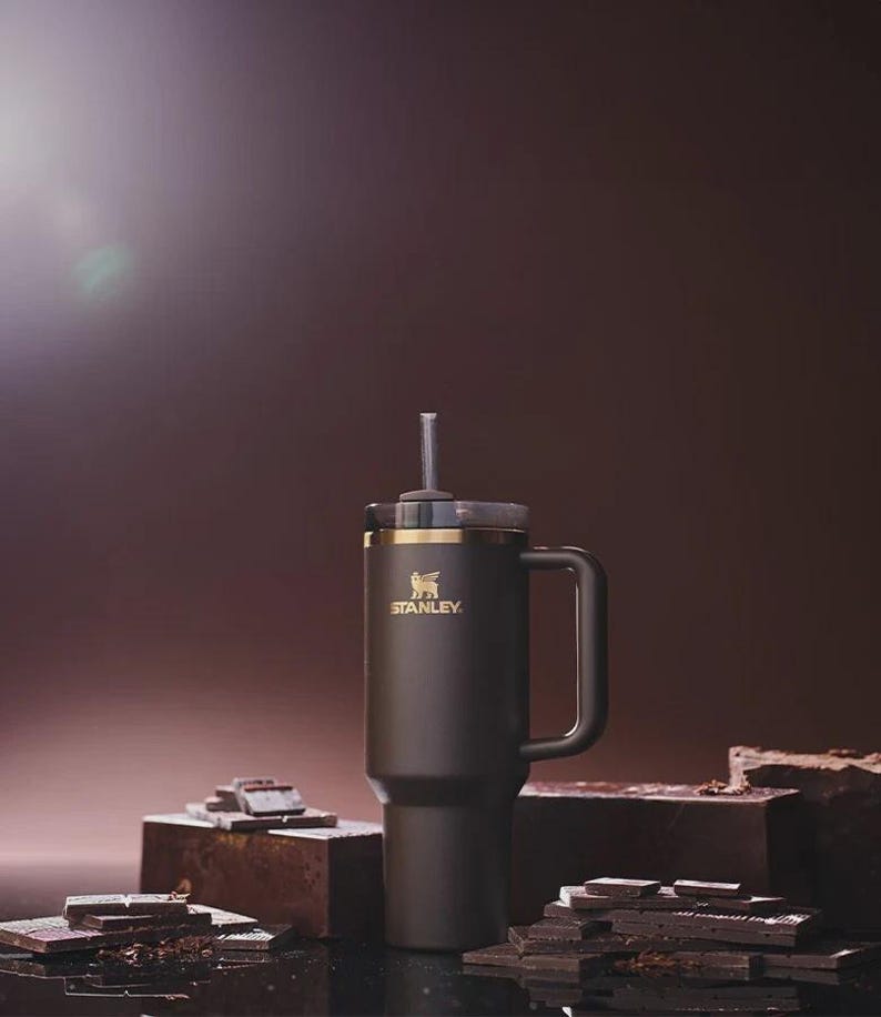Peut inclure: Un mug de voyage Stanley noir avec une poign&eacute;e et une paille. Le mug a un bord dor&eacute; et le logo Stanley en or. Le mug est pos&eacute; sur une surface sombre avec des morceaux de chocolat.