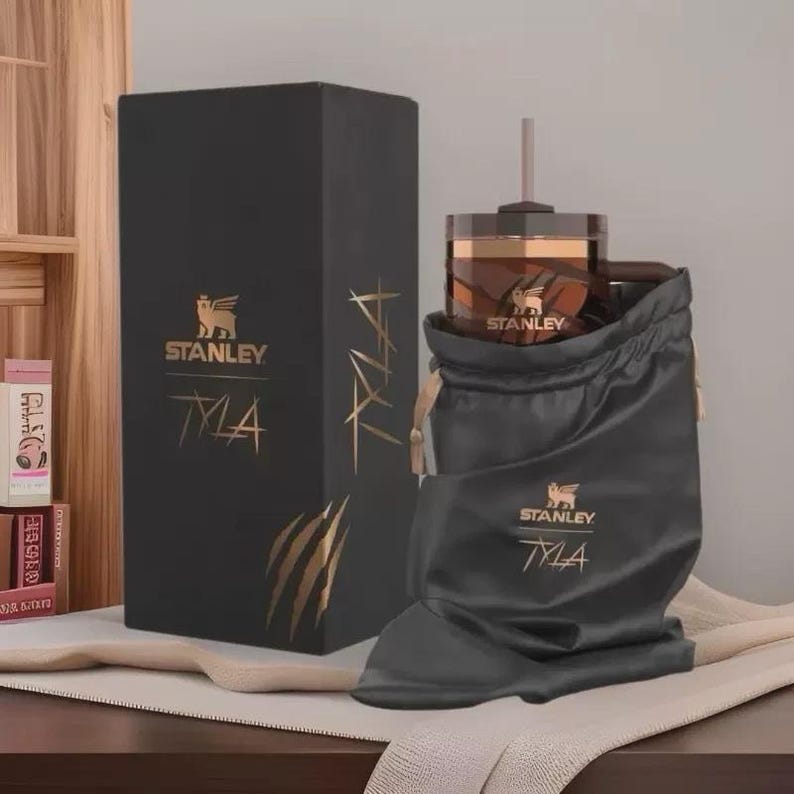 May include: A black gift set with a Stanley tumbler and a drawstring bag. The tumbler is brown and gold, with a straw. The box and bag have the Stanley logo and the text "TYLA" in gold. The set is displayed on a wooden surface.