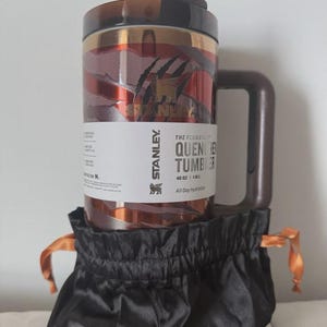 May include: A Stanley Quencher tumbler with a brown handle and lid, featuring a red and gold marbled design. The tumbler is labeled "THE FLOWSTATE QUENCHER TUMBLER" and is in a black drawstring bag with orange ties. It holds 1.18L.