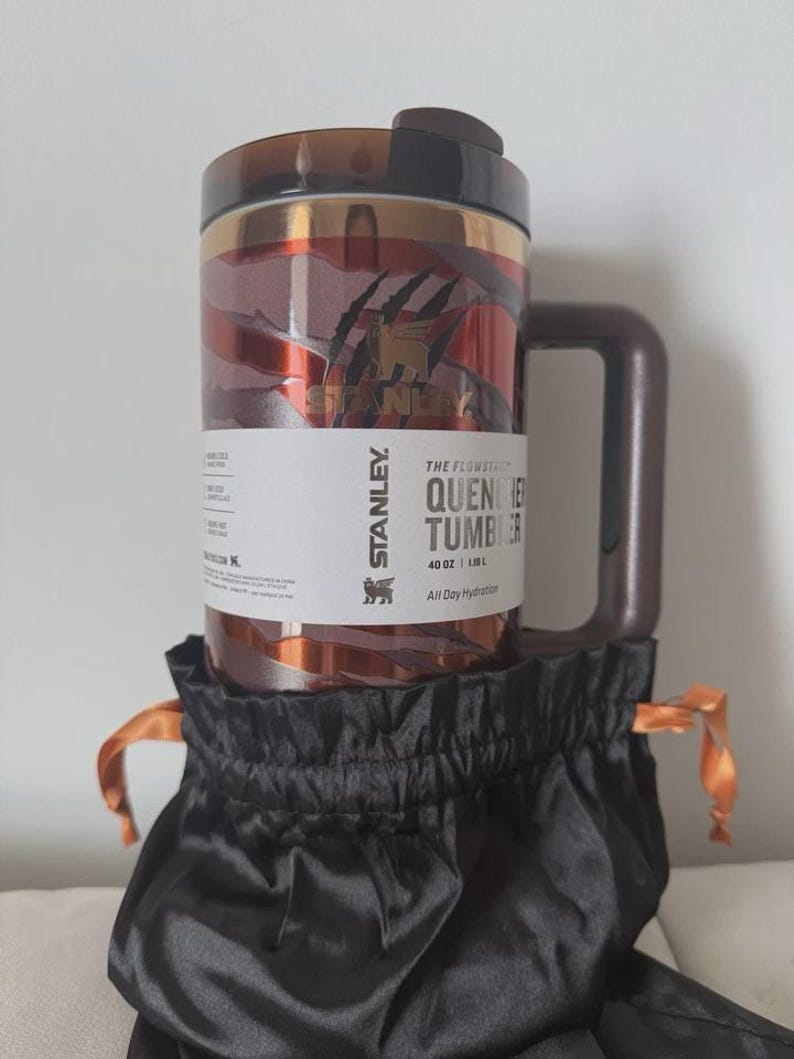 May include: A Stanley Quencher tumbler with a brown handle and lid, featuring a red and gold marbled design. The tumbler is labeled "THE FLOWSTATE QUENCHER TUMBLER" and is in a black drawstring bag with orange ties. It holds 1.18L.