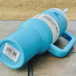 May include: A light blue Stanley Quencher H2.0 tumbler with a white band and handle. The tumbler has a straw and a handle for easy carrying. The text "STANLEY QUENCHER H2.0" is printed on the side.