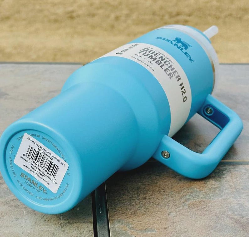 May include: A light blue Stanley Quencher H2.0 tumbler with a white band and handle. The tumbler has a straw and a handle for easy carrying. The text "STANLEY QUENCHER H2.0" is printed on the side.