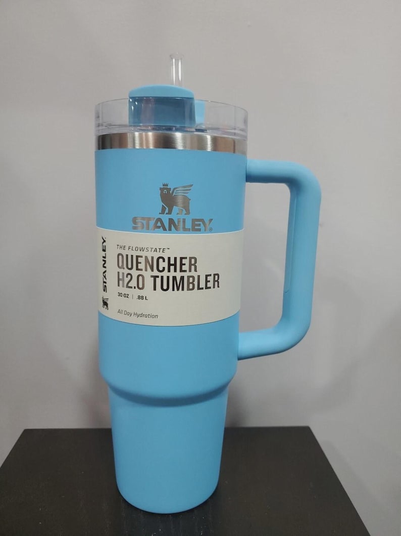 May include: A light blue Stanley Quencher H2.0 Tumbler with a handle and straw. The tumbler has a clear lid and a white label with the text "QUENCHER H2.0 TUMBLER" and "30 OZ | .88 L". The Stanley logo is visible on the front.