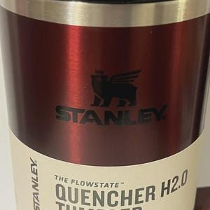 May include: A red Stanley Quencher H2.0 tumbler. The tumbler has a black lid and a silver band. The Stanley logo is displayed in black. The label reads "QUENCHER H2.0 TUMBLER" and "40 OZ | 1.18 L".