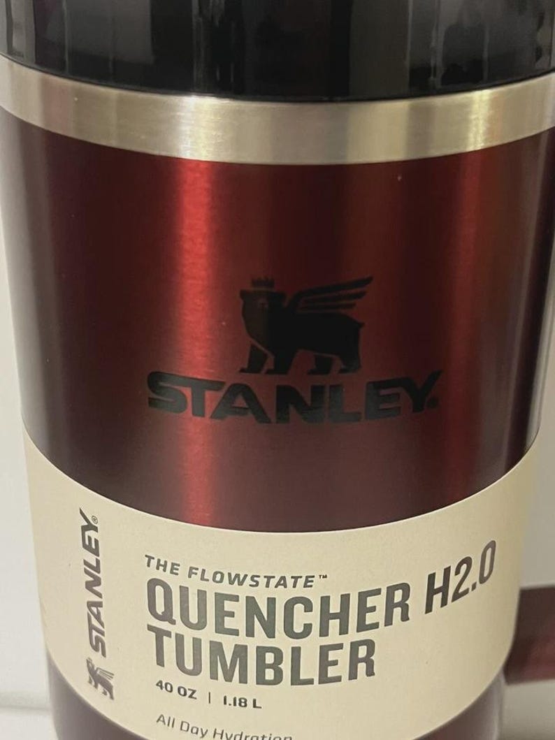 May include: A red Stanley Quencher H2.0 tumbler. The tumbler has a black lid and a silver band. The Stanley logo is displayed in black. The label reads "QUENCHER H2.0 TUMBLER" and "40 OZ | 1.18 L".