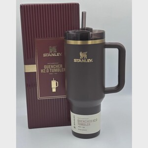 Peut inclure: Un gobelet Stanley Quencher H2.0 marron fonc&eacute; avec une poign&eacute;e et une paille, &agrave; c&ocirc;t&eacute; d'une bo&icirc;te bordeaux assortie. Le gobelet a un bord dor&eacute; et le logo Stanley. La bo&icirc;te a des lettres dor&eacute;es et un logo assorti.
