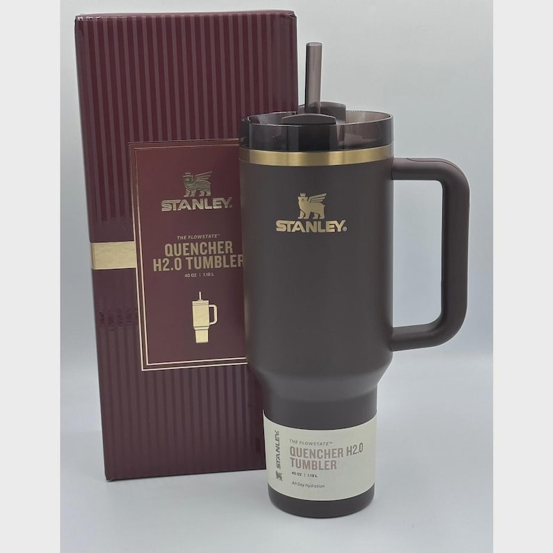 Peut inclure: Un gobelet Stanley Quencher H2.0 marron fonc&eacute; avec une poign&eacute;e et une paille, &agrave; c&ocirc;t&eacute; d'une bo&icirc;te bordeaux assortie. Le gobelet a un bord dor&eacute; et le logo Stanley. La bo&icirc;te a des lettres dor&eacute;es et un logo assorti.