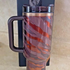 May include: A stainless steel travel mug with a handle, featuring a brown and copper tiger stripe design. The mug has a copper-coloured rim and a dark brown handle. A black box is behind the mug.