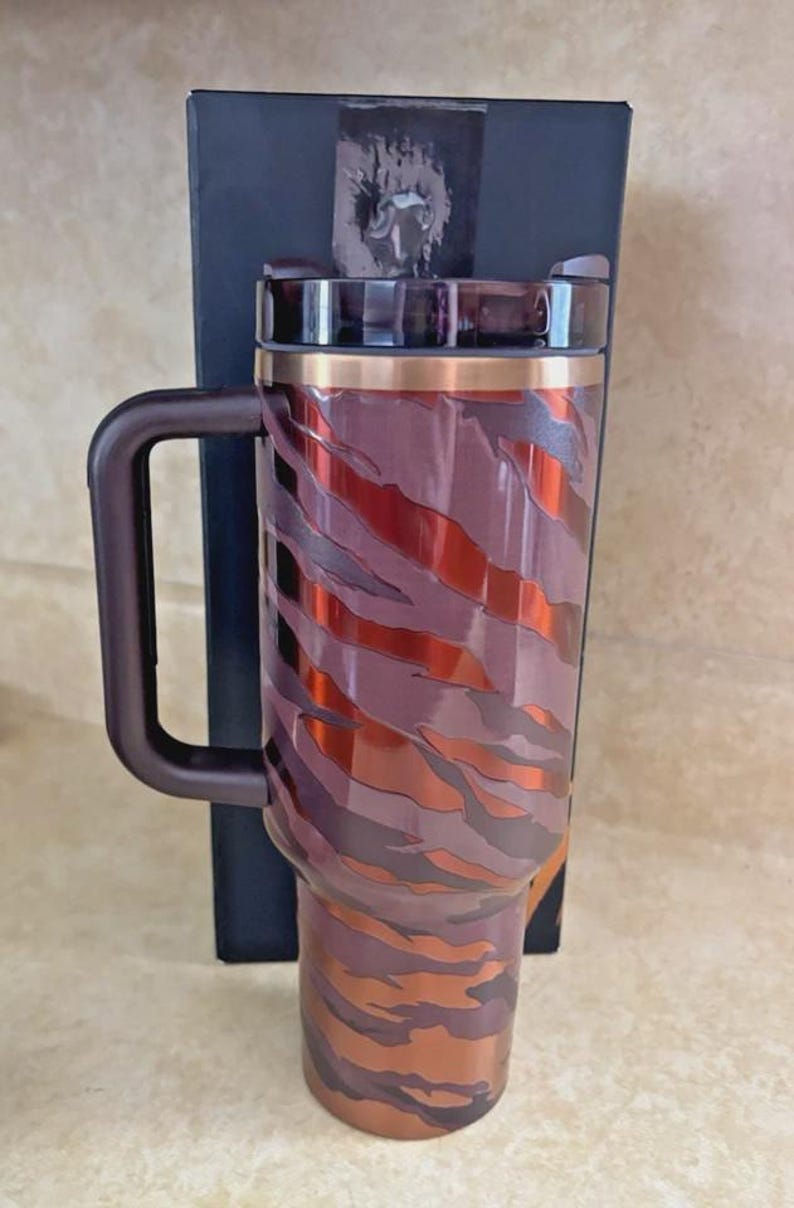 May include: A stainless steel travel mug with a handle, featuring a brown and copper tiger stripe design. The mug has a copper-coloured rim and a dark brown handle. A black box is behind the mug.