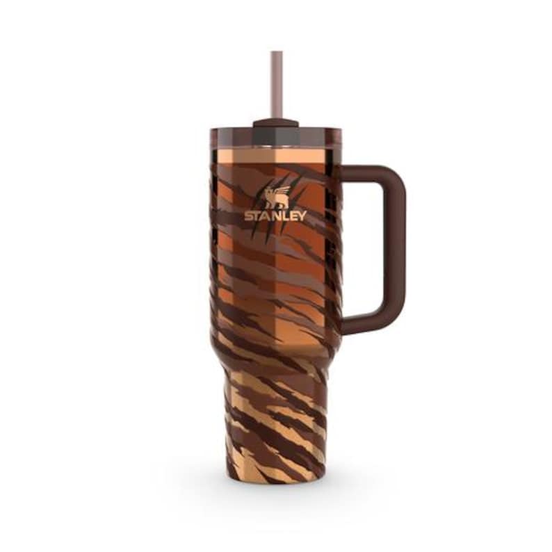 May include: A Stanley insulated tumbler with a handle and straw. The tumbler has a brown and tan tiger stripe pattern. The Stanley logo is visible on the front. Designed to keep drinks cold or hot. Holds approximately 946 ml.
