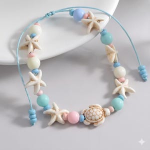 May include: A light blue adjustable bracelet featuring starfish, turtle, and round beads in pastel colors. The bracelet has a nautical theme, with white, pink, and blue accents. The bracelet is made of a light blue cord.