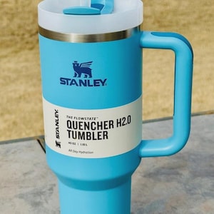 May include: A light blue Stanley Quencher H2.0 tumbler with a white lid and handle. The tumbler has a straw and the text "THE FLOWSTATE QUENCHER H2.0 TUMBLER" printed on the side. It holds 1.18L and is designed for all-day hydration.