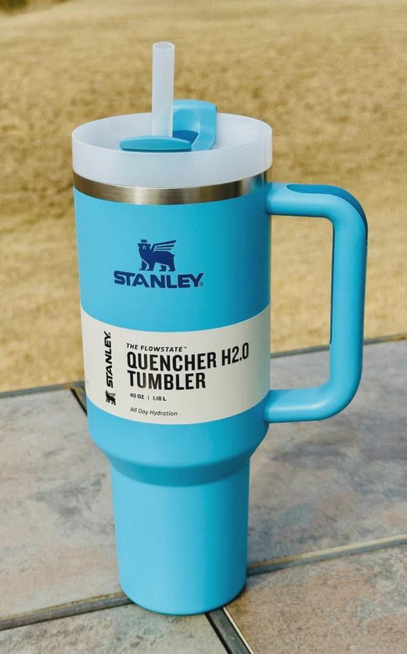 May include: A light blue Stanley Quencher H2.0 tumbler with a white lid and handle. The tumbler has a straw and the text "THE FLOWSTATE QUENCHER H2.0 TUMBLER" printed on the side. It holds 1.18L and is designed for all-day hydration.
