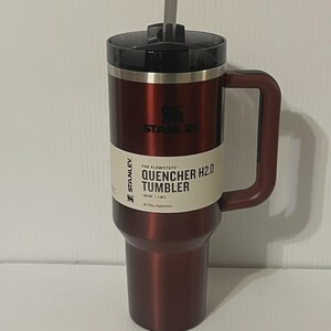 May include: A maroon insulated tumbler with a black lid and handle. The tumbler has a straw and a white label with the text "QUENCHER H2.0 TUMBLER" and "40 OZ | 1.18 L". The brand name "STANLEY" is visible.