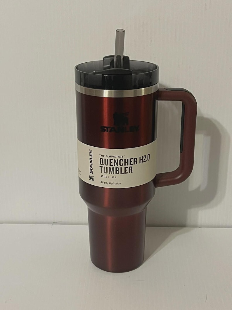 May include: A maroon insulated tumbler with a black lid and handle. The tumbler has a straw and a white label with the text "QUENCHER H2.0 TUMBLER" and "40 OZ | 1.18 L". The brand name "STANLEY" is visible.