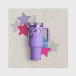 Op de afbeelding: Een lavendelkleurige Stanley-beker met een handvat, deksel en rietje. De beker is versierd met sterren en strikken. Het Stanley-logo is zichtbaar. De achtergrond heeft steruitsparingen in verschillende kleuren.