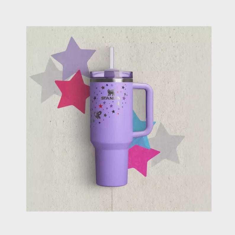 Op de afbeelding: Een lavendelkleurige Stanley-beker met een handvat, deksel en rietje. De beker is versierd met sterren en strikken. Het Stanley-logo is zichtbaar. De achtergrond heeft steruitsparingen in verschillende kleuren.