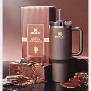 Peut inclure: Un gobelet Stanley Quencher H2.0 marron avec une poign&eacute;e et une paille, &agrave; c&ocirc;t&eacute; d'une bo&icirc;te assortie. La bo&icirc;te porte le logo Stanley et des informations sur le produit. Le gobelet est pr&eacute;sent&eacute; avec des morceaux de chocolat.