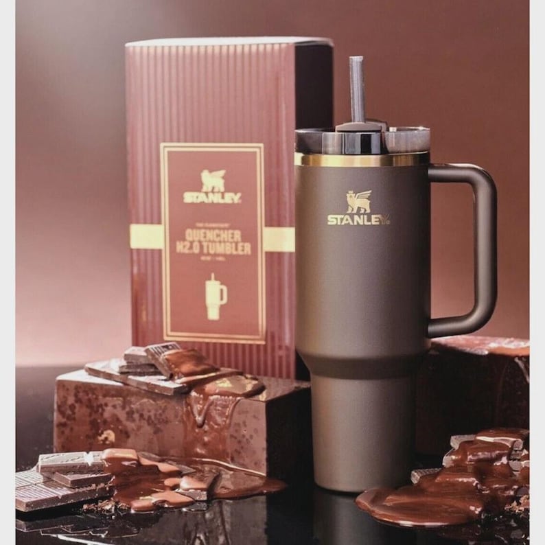 Peut inclure: Un gobelet Stanley Quencher H2.0 marron avec une poign&eacute;e et une paille, &agrave; c&ocirc;t&eacute; d'une bo&icirc;te assortie. La bo&icirc;te porte le logo Stanley et des informations sur le produit. Le gobelet est pr&eacute;sent&eacute; avec des morceaux de chocolat.