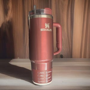 May include: A red Stanley insulated travel mug with a handle and a straw. The mug has the Stanley logo and icons on the bottom. The mug is designed to keep beverages hot or cold for extended periods.