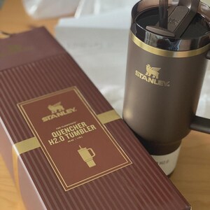 Peut inclure: Un gobelet Stanley Quencher H2.0 marron avec une poign&eacute;e et une bo&icirc;te marron assortie. La bo&icirc;te porte des lettres dor&eacute;es et un bord dor&eacute;, avec les mots "The Flowstate Quencher H2.0 Tumbler" et un dessin du gobelet. Le gobelet contient 1,18L.