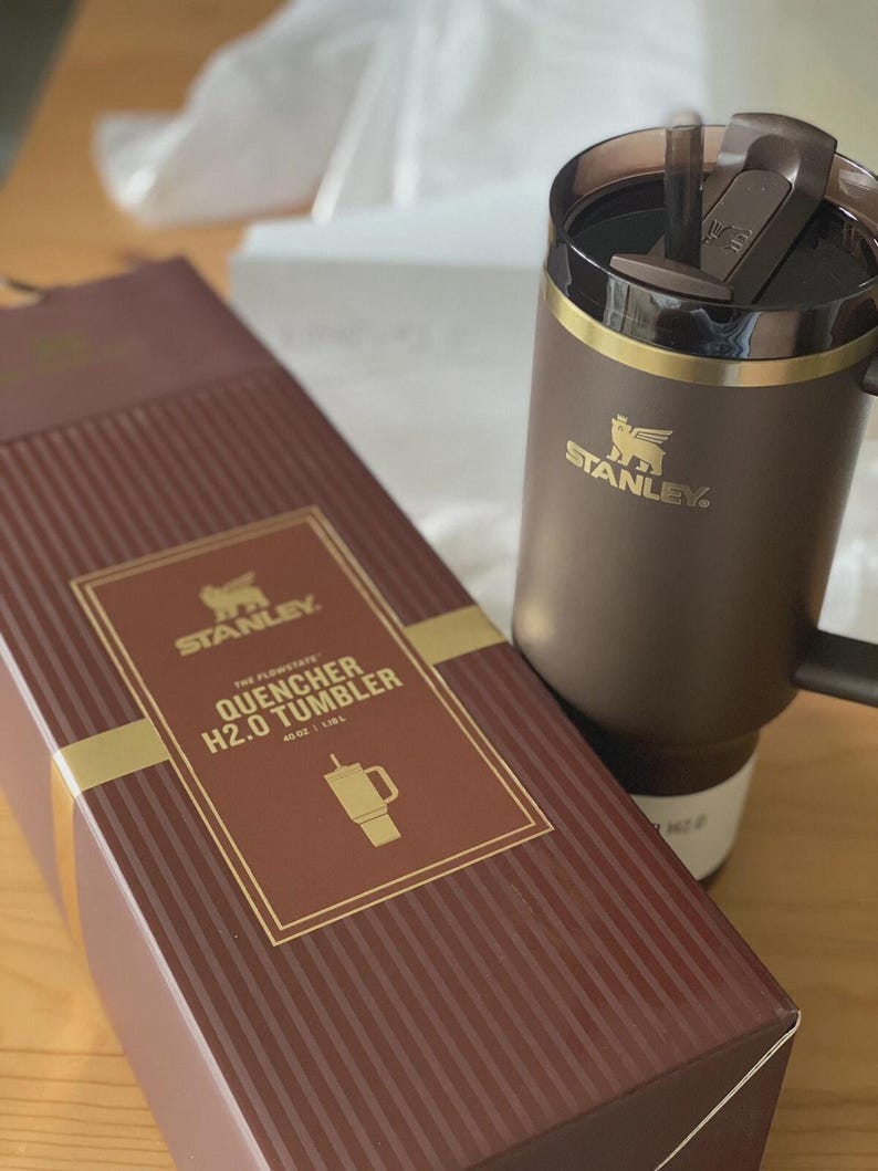 Peut inclure: Un gobelet Stanley Quencher H2.0 marron avec une poign&eacute;e et une bo&icirc;te marron assortie. La bo&icirc;te porte des lettres dor&eacute;es et un bord dor&eacute;, avec les mots "The Flowstate Quencher H2.0 Tumbler" et un dessin du gobelet. Le gobelet contient 1,18L.