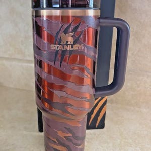 May include: A Stanley travel mug with a tiger stripe pattern in brown, orange, and purple. The mug has a dark purple handle and lid. The Stanley logo is visible. A black box with tiger stripes is behind the mug. The mug is made of stainless steel.