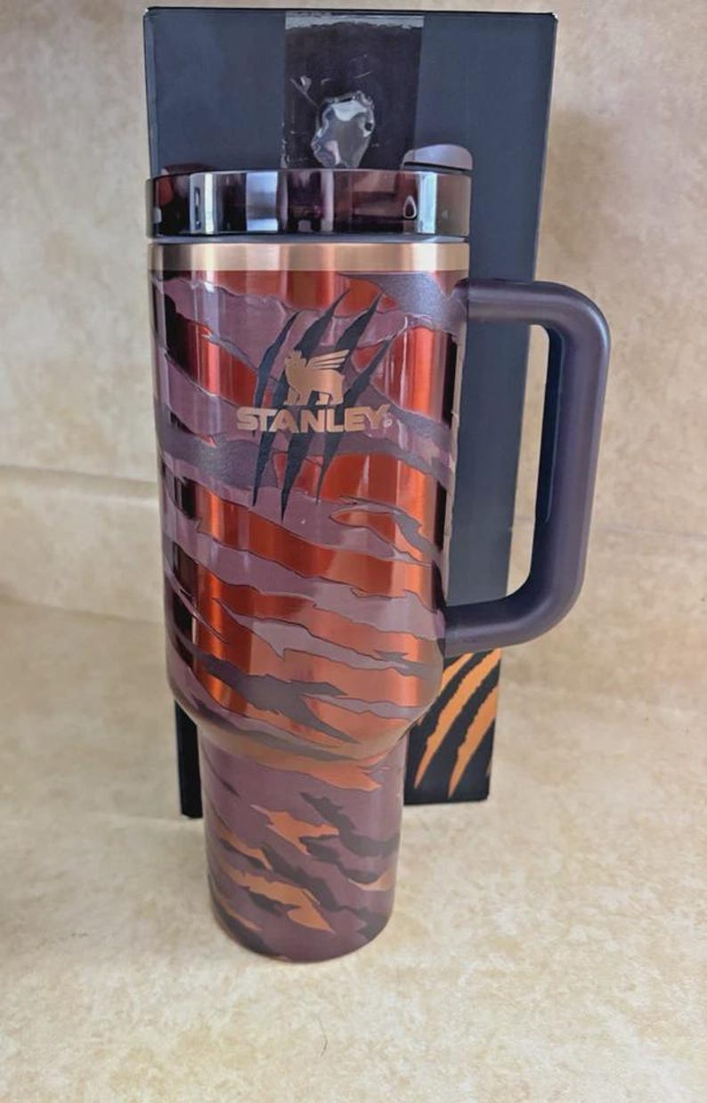 May include: A Stanley travel mug with a tiger stripe pattern in brown, orange, and purple. The mug has a dark purple handle and lid. The Stanley logo is visible. A black box with tiger stripes is behind the mug. The mug is made of stainless steel.