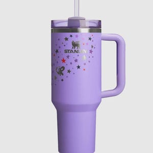 Op de afbeelding: Een lavendelkleurige Stanley-beker met een handvat en een rietje. De beker heeft een zilveren logo en ster- en vlindergraphics. Het deksel is transparant en het handvat heeft dezelfde lavendelkleur als de beker.
