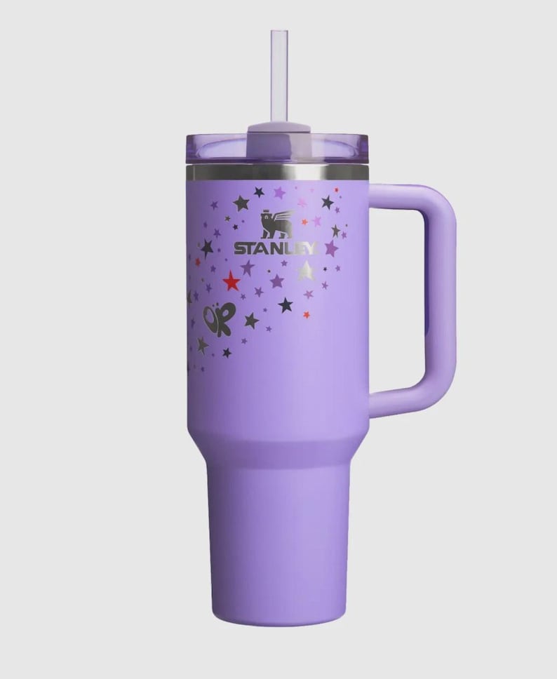 Op de afbeelding: Een lavendelkleurige Stanley-beker met een handvat en een rietje. De beker heeft een zilveren logo en ster- en vlindergraphics. Het deksel is transparant en het handvat heeft dezelfde lavendelkleur als de beker.