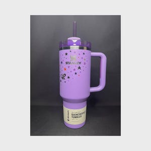 Op de afbeelding: Een paarse Stanley Quencher H2.0 FlowState tumbler met een handvat en een rietje. De tumbler heeft een sterrenbeeldontwerp met sterren en het Stanley-logo. De tekst "OR" is ook zichtbaar.