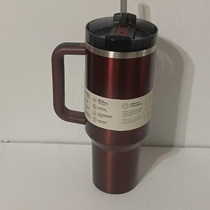 May include: A maroon insulated tumbler with a handle and a black lid. The tumbler has a silver rim and a matching maroon handle. A gray straw extends from the lid. The tumbler has a white label with text.
