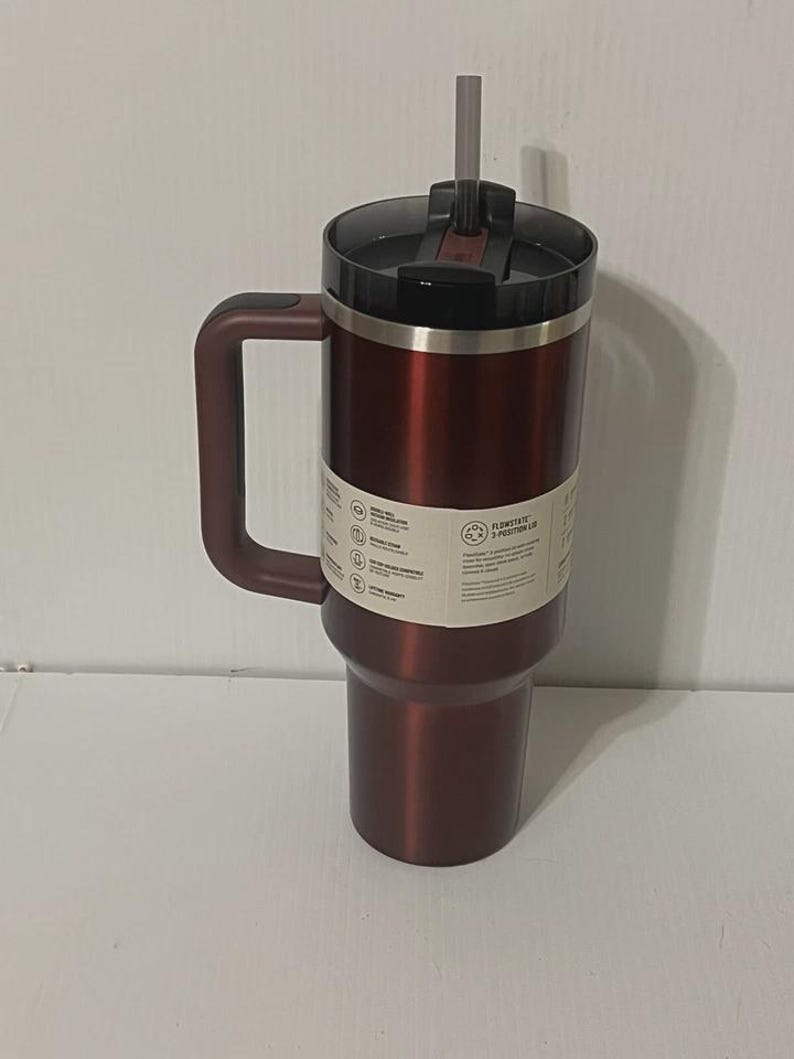 May include: A maroon insulated tumbler with a handle and a black lid. The tumbler has a silver rim and a matching maroon handle. A gray straw extends from the lid. The tumbler has a white label with text.