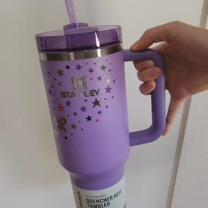 Op de afbeelding: Een paarse Stanley Quencher H2.0 tumbler met een handvat en een rietje. De tumbler heeft een deksel en is versierd met sterrenafbeeldingen. De tekst "STANLEY" is zichtbaar op de zijkant. De tumbler is ontworpen voor de hele dag hydratatie.