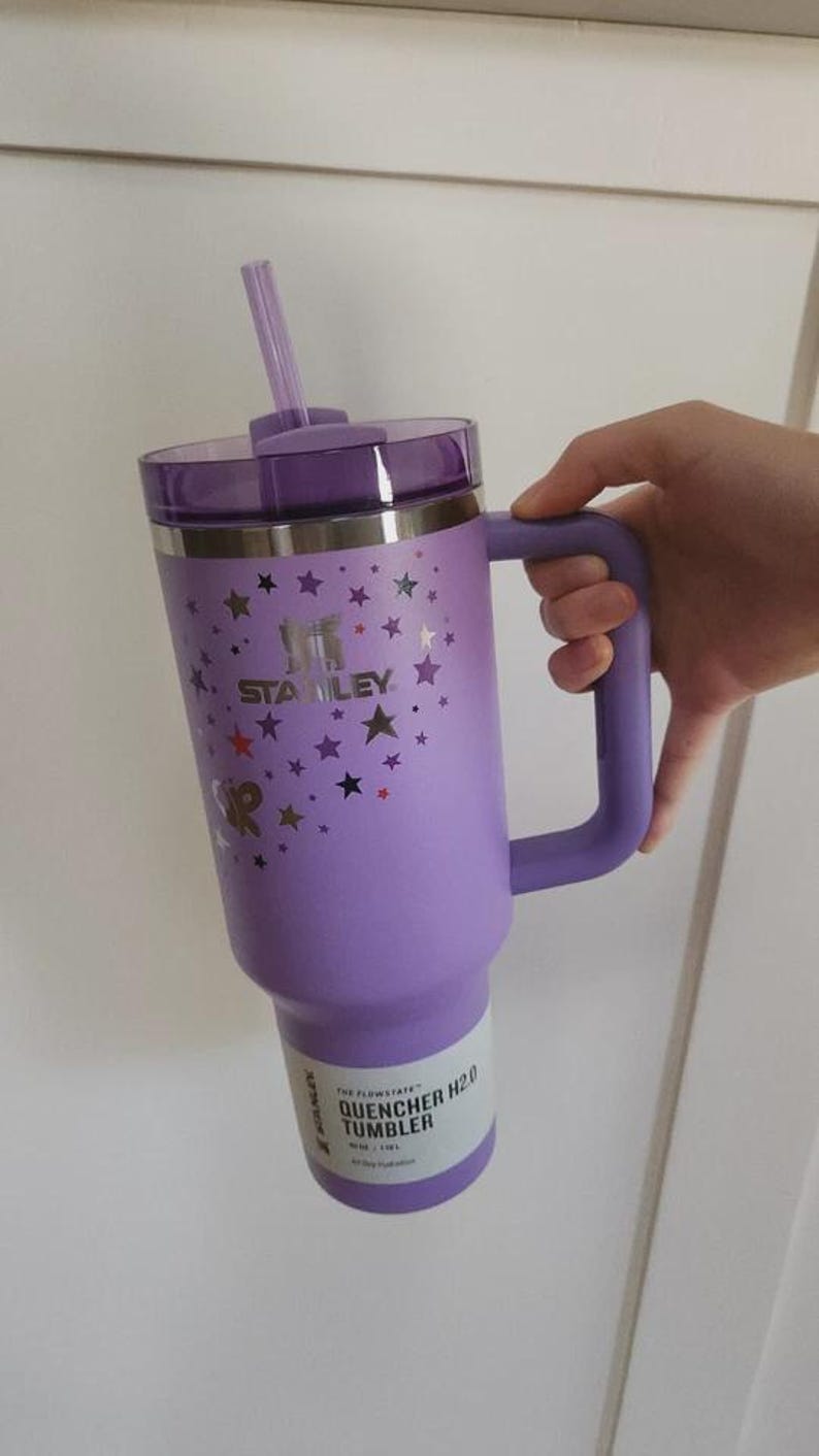 Op de afbeelding: Een paarse Stanley Quencher H2.0 tumbler met een handvat en een rietje. De tumbler heeft een deksel en is versierd met sterrenafbeeldingen. De tekst "STANLEY" is zichtbaar op de zijkant. De tumbler is ontworpen voor de hele dag hydratatie.