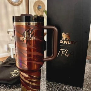 May include: A brown and gold Stanley travel mug with a handle and straw. The mug features a tiger stripe design and the Stanley logo. A black box with the Stanley logo and the text "M4" is next to the mug. A small black bag with the Stanley logo is also present.