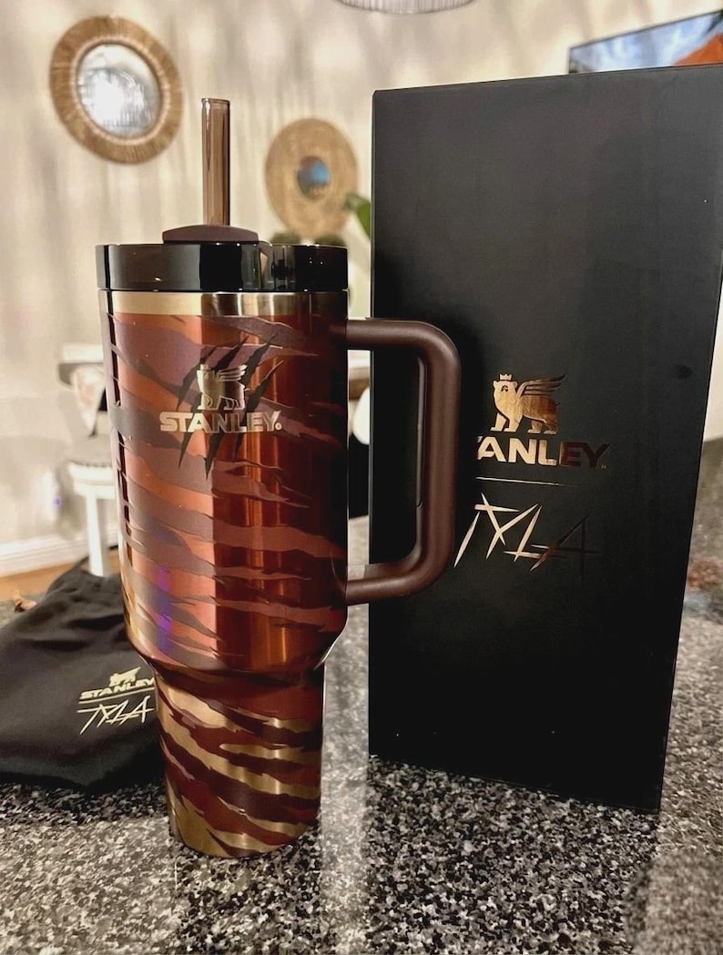May include: A brown and gold Stanley travel mug with a handle and straw. The mug features a tiger stripe design and the Stanley logo. A black box with the Stanley logo and the text "M4" is next to the mug. A small black bag with the Stanley logo is also present.