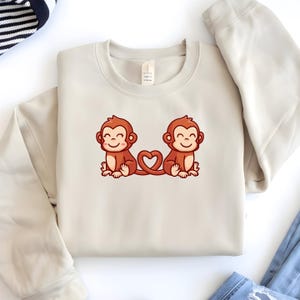 May include: Beige sweatshirt featuring a cartoon illustration of two smiling monkeys with a heart-shaped tail. The monkeys are brown with pink cheeks and are sitting side by side. The sweatshirt is a neutral color and has a crew neck.