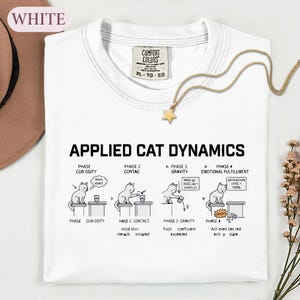 May include: A white t-shirt featuring a graphic titled APPLIED CAT DYNAMICS, illustrating four phases of a cat knocking an object off a table. The design includes line drawings of a cat, text labels for each phase, and a gold star pendant necklace.