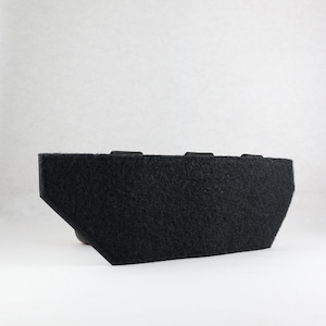 May include: A black, rectangular object with a felt-like texture. The object has a trapezoidal shape on one side and two small rectangular protrusions on the top. The background is plain white.