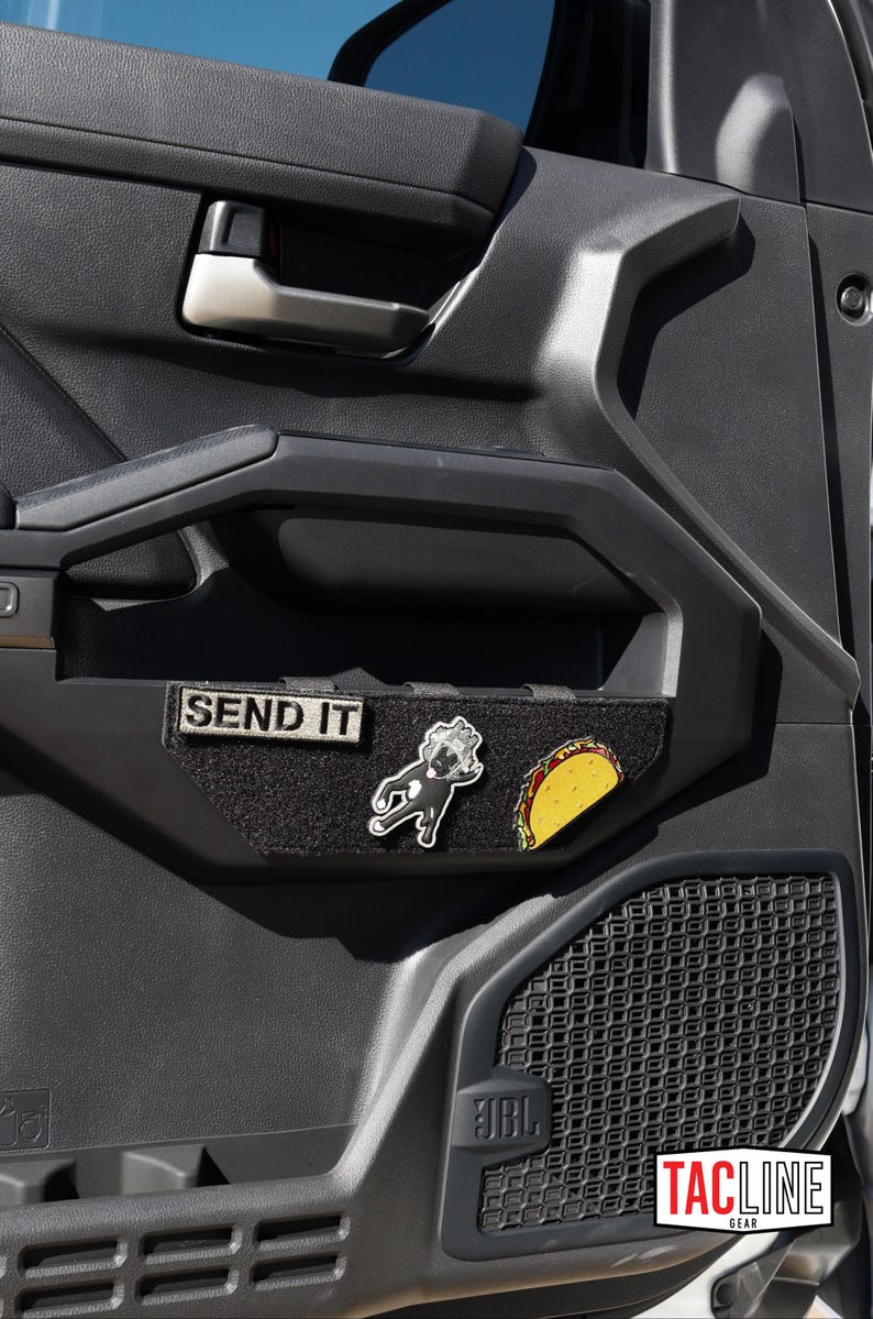 May include: Black interior door panel with a silver handle and a black fabric patch that reads "SEND IT." The patch has two enamel pins: a cartoon dog and a taco. A JBL speaker is visible.