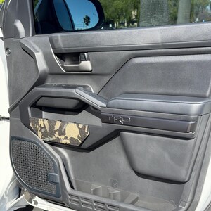 May include: A close-up of a car door interior. The door is black with a gray armrest and a camouflage-patterned pocket. The door has a TRD logo and a speaker grill. The window is tinted, and the side mirror is visible.