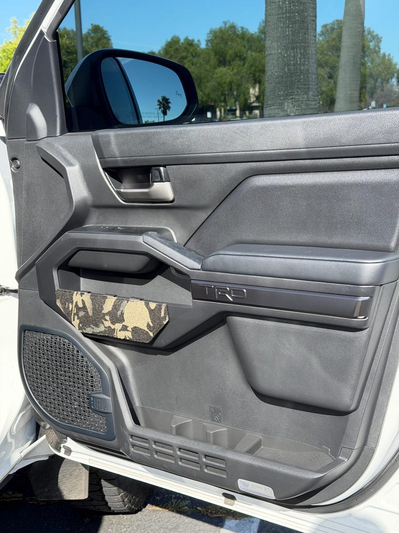 May include: A close-up of a car door interior. The door is black with a gray armrest and a camouflage-patterned pocket. The door has a TRD logo and a speaker grill. The window is tinted, and the side mirror is visible.