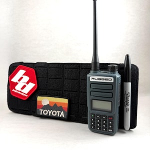 May include: A gray and black handheld radio with the word "RUGGED" printed on it, a black Sharpie marker, and a black pouch with patches that say "bd bajadoslans" and "TOYOTA".