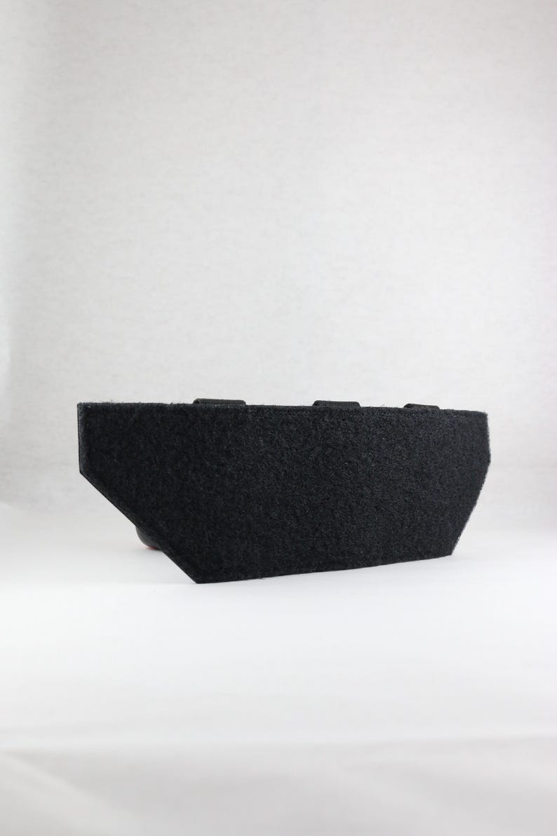 May include: A black, rectangular object with a felt-like texture. The object has a trapezoidal shape on one side and two small rectangular protrusions on the top. The background is plain white.