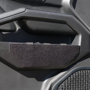 May include: Black interior door panel with a textured surface. A black, rectangular, fabric-covered insert is placed in the door handle area. The JBL and TACLINE GEAR logos are visible.
